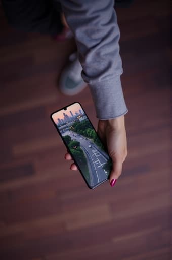 한 사람이 손에 세련된 스마트폰을 들고 있으며, 고해상도 디스플레이가 강조되고 있습니다. 배경은 흐릿한 도시 풍경으로, 역동적인 도시 생활을 암시하며, 녹지와 현대 건축의 느낌이 있습니다. 전체적인 분위기는 생동감 있고 현대적이며, 기술 중심의 라이프스타일을 반영합니다.