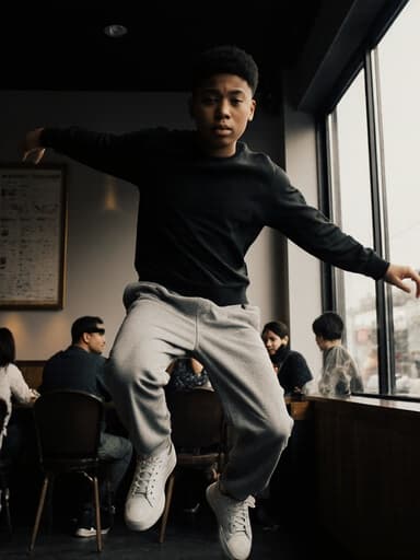 한 젊은 혼혈 남성이 현대적인 카페에서 편안한 운동복과 스니커즈를 입고 점프하고 있습니다. 그의 약간 의기소침한 표정은 앉아 있는 손님들로 가득한 카페의 활기찬 배경과 대조를 이루어, 에너지와 내면의 성찰을 모두 담고 있는 장면을 만들어냅니다.