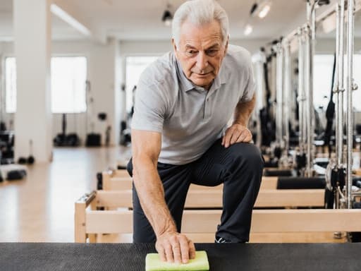 친근한 60대의 백인 남성이 밝고 깔끔한 필라테스 스튜디오에서 필라테스 매트를 청소하며 따뜻하게 미소를 짓고 있습니다. 그는 편안한 필라테스 복장과 운동화를 착용하고 있으며, 청소 도구를 손에 들고 있습니다. 스튜디오는 다양한 필라테스 기구로 가득 차 있어 건강과 웰빙을 촉진하는 매력적인 분위기를 만들어냅니다.