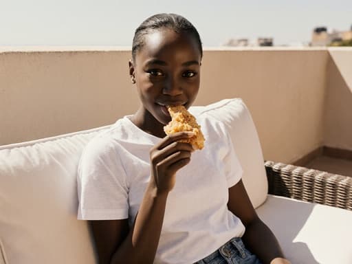 10대 초반의 아프리카계 소녀가 캐주얼한 티셔츠와 청바지를 입고 햇살이 비치는 테라스에 앉아 음식을 들고 미소를 지으며 즐기는 모습입니다. 따뜻한 분위기가 편안함과 즐거움을 자아냅니다.