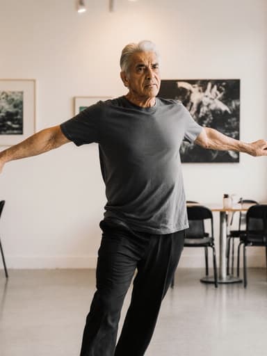 60대 라틴계 남성이 편안한 착용의 티셔츠와 바지를 입고 춤을 연습하는 모습의 풀 바디 샷. 그는 팔을 약간 벌리고 다리를 힘 있게 고정한 자세로 서 있으며, 집중한 표정으로 긴장감을 나타냅니다. 배경은 부드러운 조명이 비추는 현대적인 카페 내부로, 벽에는 다양한 예술 작품이 걸려 있어 그의 춤 연습과 잘 어울리는 창의적인 분위기를 더하고 있습니다.